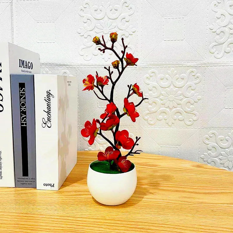 Artificial plum blossom plant with red flowers in a small white pot, set on a wooden desk next to decorative books.