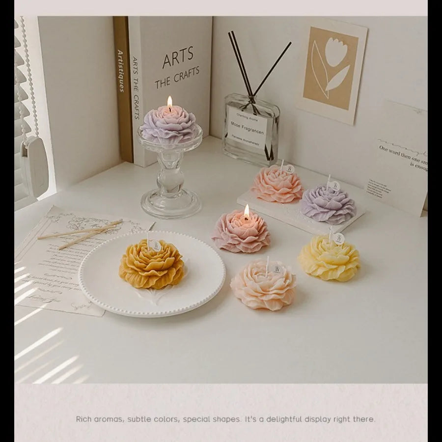 Assorted pastel-coloured flower-shaped decorative candles arranged on a white table with a glass candle holder.