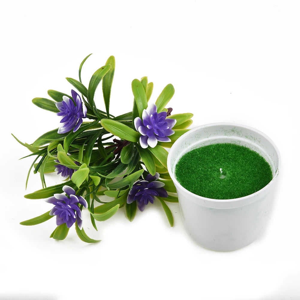 Artificial purple flowers with green leaves next to a white pot filled with faux green grass on a white background.