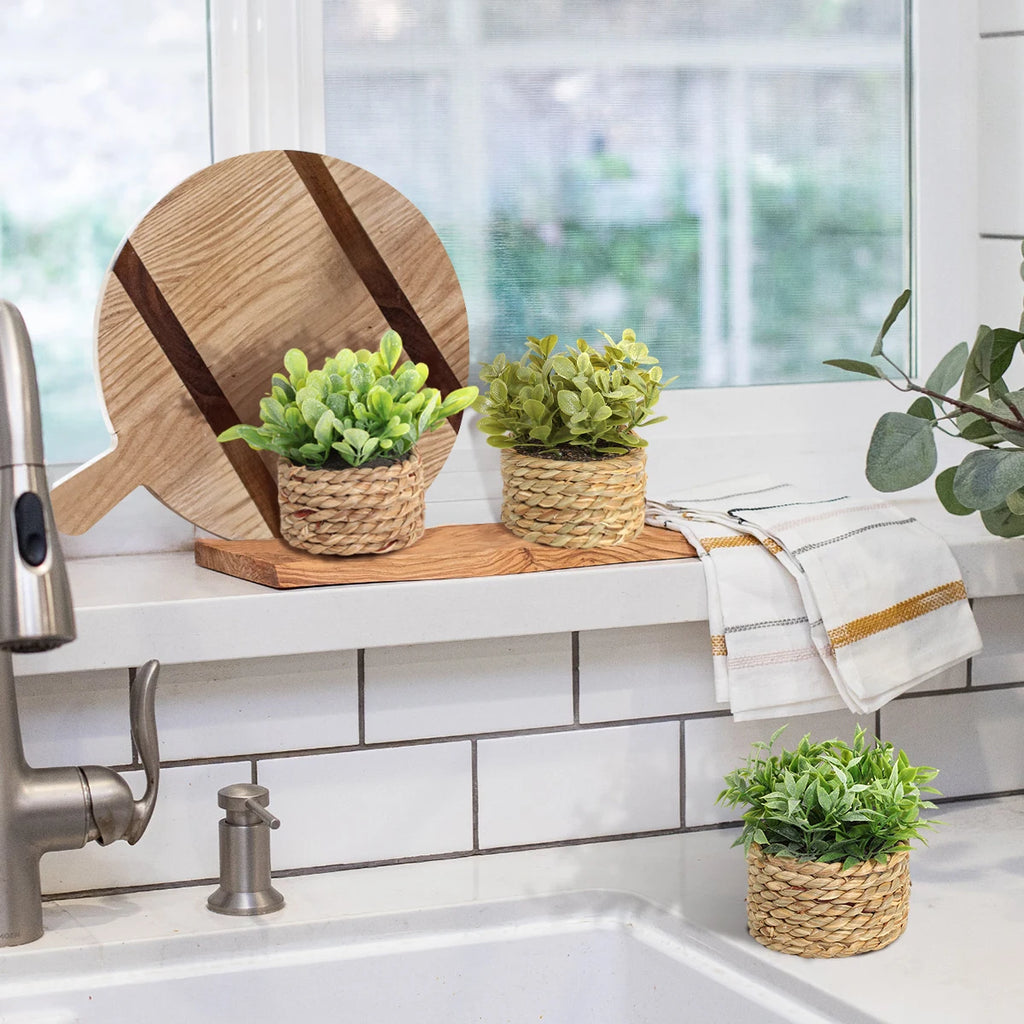 Three small artificial green plants in woven grass pots displayed on a kitchen sink ledge with wooden decor.