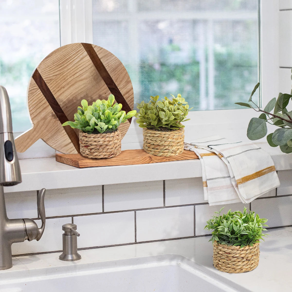 Three small artificial green plants in woven grass pots displayed on a kitchen sink ledge with wooden decor.