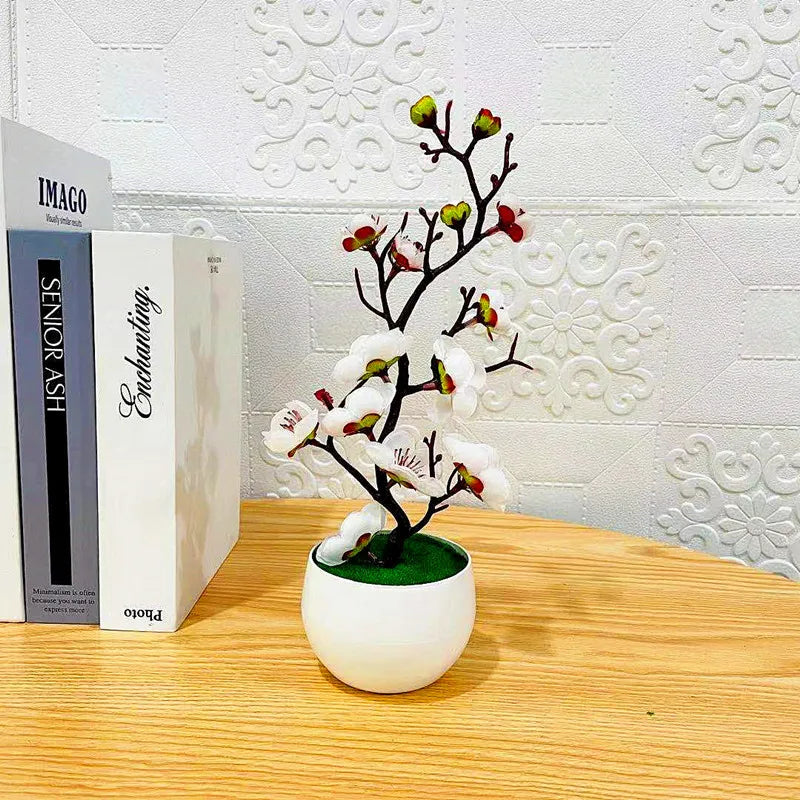 Artificial plum blossom plant in a small white pot, sitting on a wooden surface next to books.