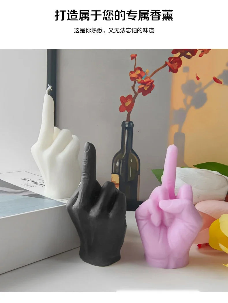 Three hand-shaped candles in white, black, and pink, each displaying a middle finger gesture on a table.