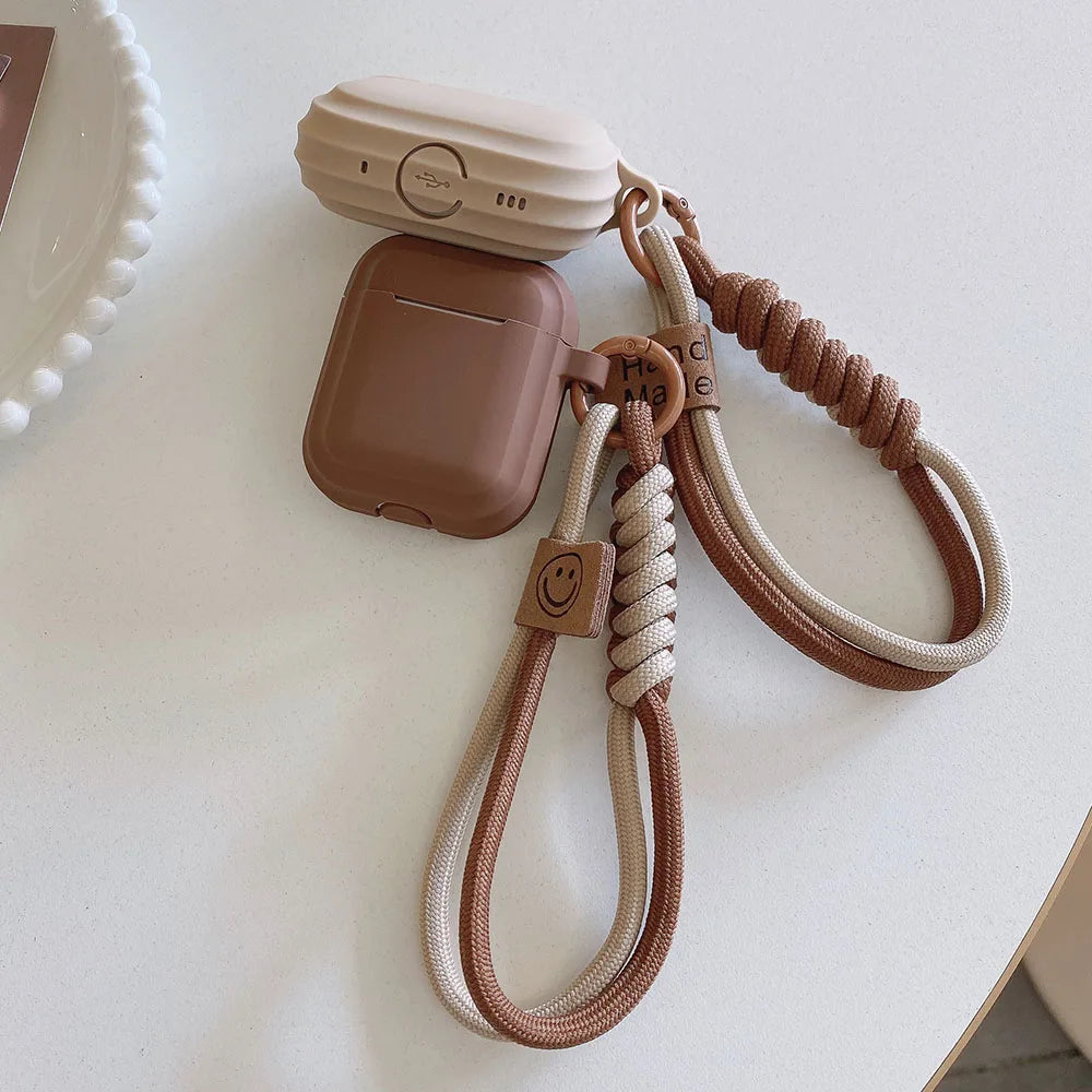 Beige and brown silicone AirPods case with a braided rope wrist strap and smiley face detail.