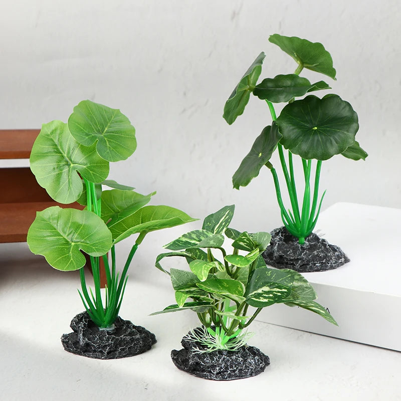 Three artificial aquarium plants with broad green leaves and sturdy black rock bases on a white surface.