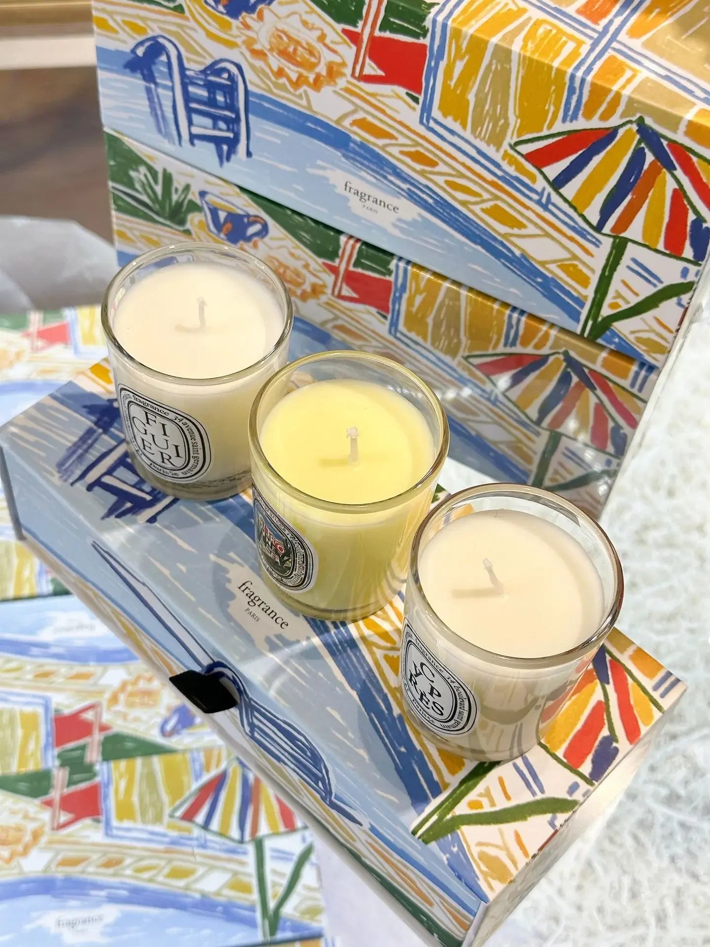 Three scented candles in glass jars on a decorative gift box with a colourful, hand-drawn seaside summer scene.