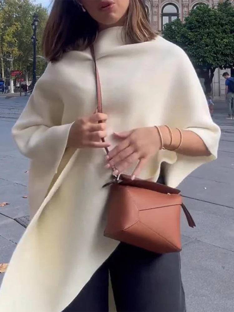 Cream knitted cape sweater with a loose, asymmetric drape and wide sleeves, worn outdoors.