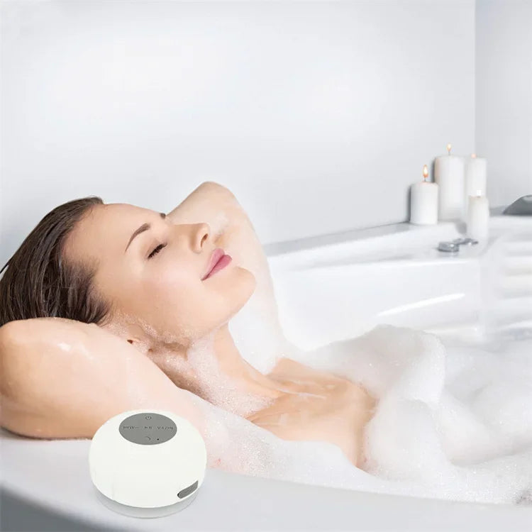 A woman relaxing in a bubbly bath next to a white, circular waterproof Bluetooth speaker with a suction cup base.