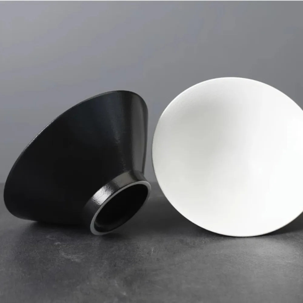 Two modern, cone-shaped bowls in matte black and white, resting on a dark grey surface. Minimalist design.