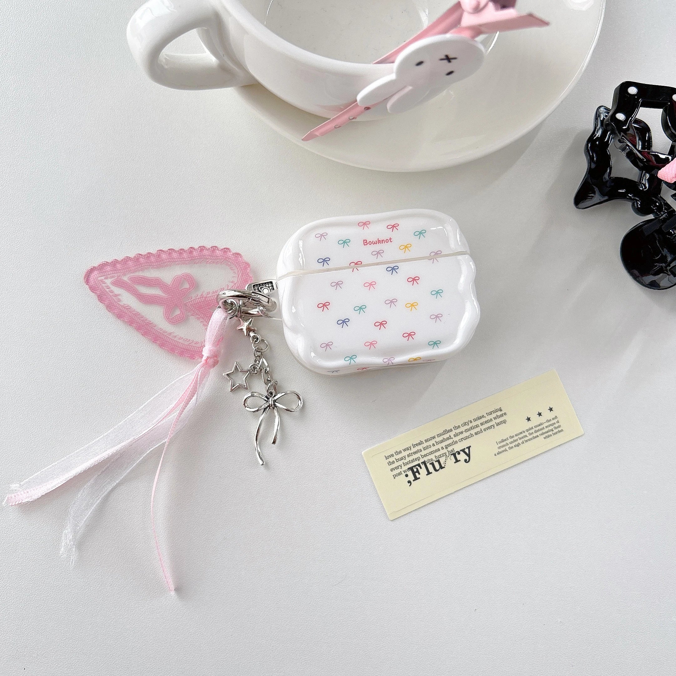 White, scalloped AirPod Pro case with colourful bow pattern, lace heart, ribbon charm and silver star keychain.