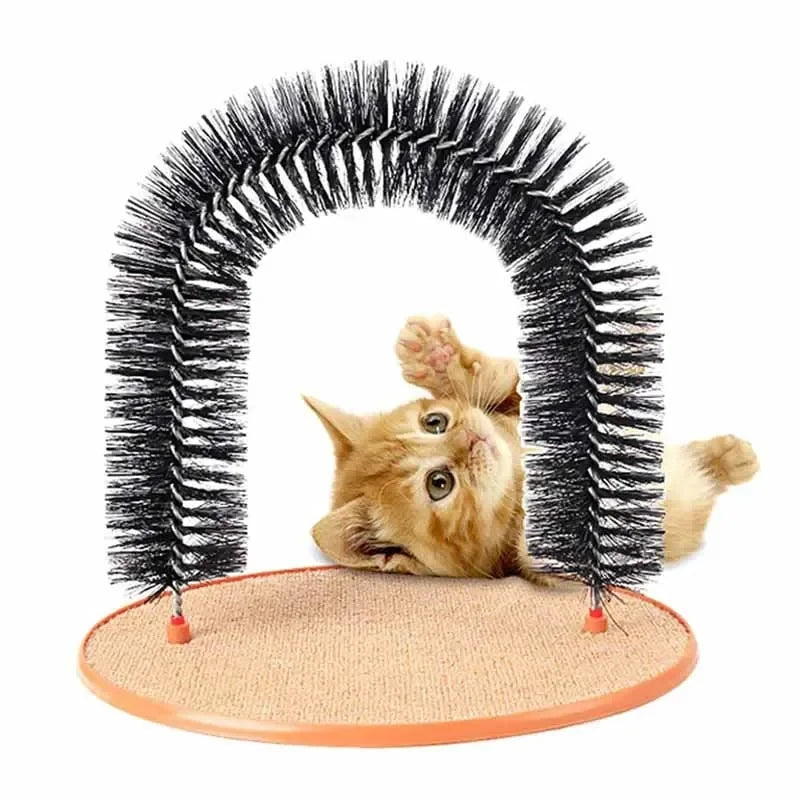 Ginger kitten playing under a black, arched cat grooming brush set on a circular, textured beige scratching pad.