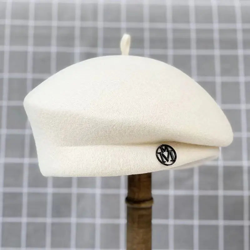 A classic cream wool felt beret with a small, circular black logo detail, shown on a mannequin head.