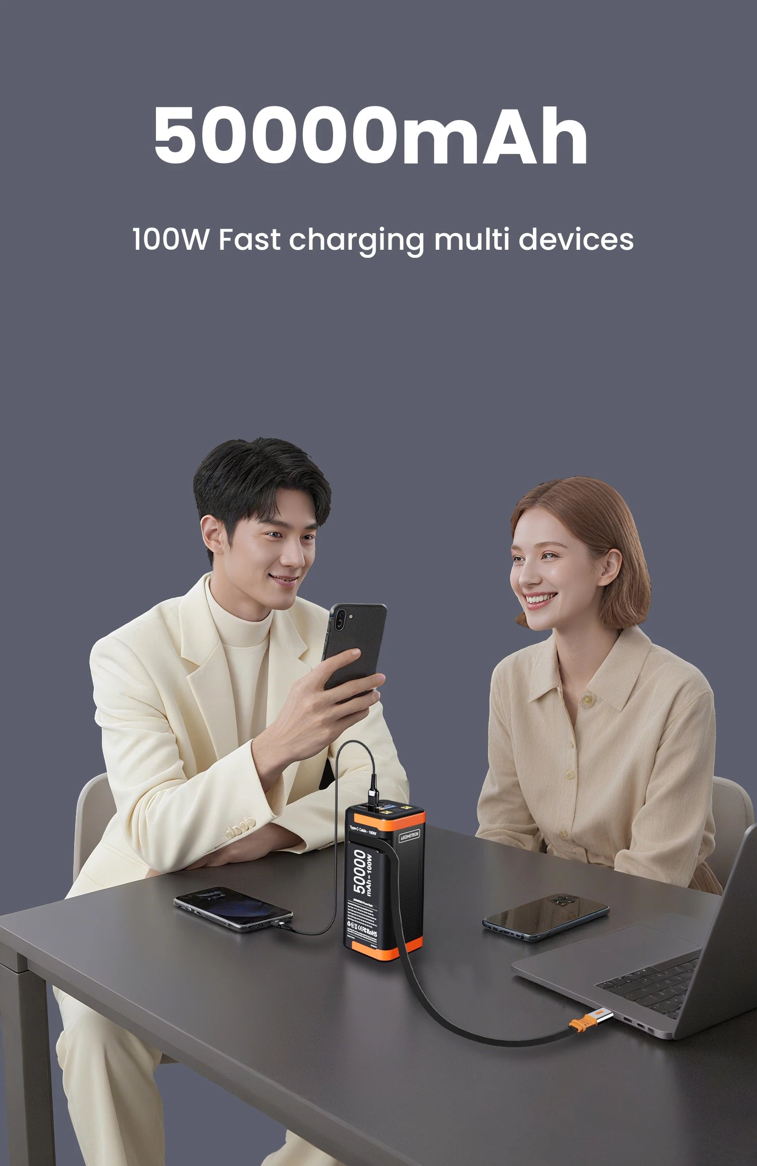 Black and orange 50000mAh portable power bank charging a laptop and smartphone simultaneously on a desk.