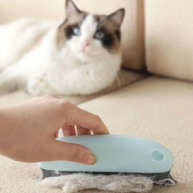 A hand using a light blue handheld lint brush to remove cat hair from a beige sofa, with a cat in the background.