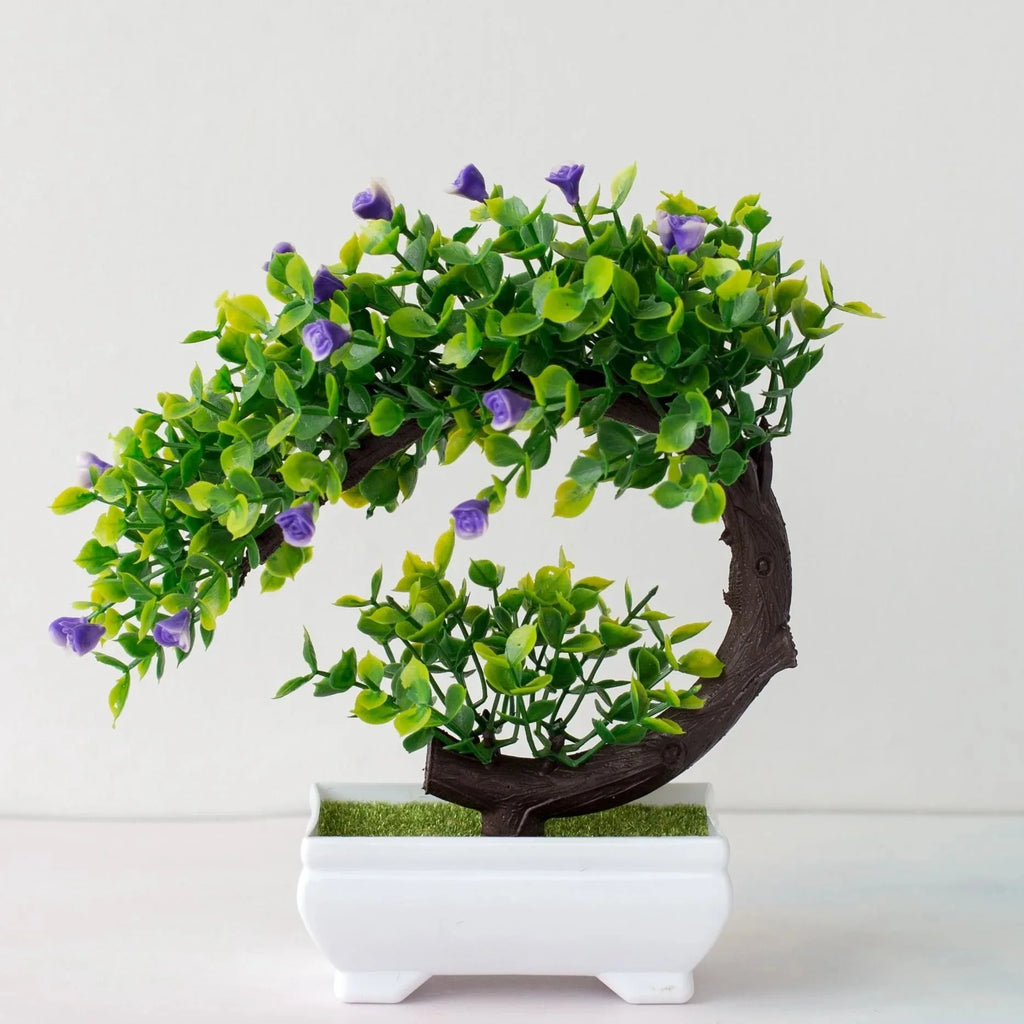Artificial bonsai tree with a curved brown trunk, green leaves, and small purple flowers in a white rectangular pot.