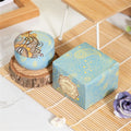Blue scented candle in a decorative patterned tin with a matching presentation box on a bamboo mat.