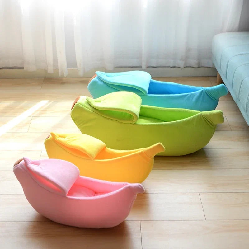 Four soft, plush banana-shaped pet beds in pink, yellow, green, and blue, featuring a peel-back hood design.