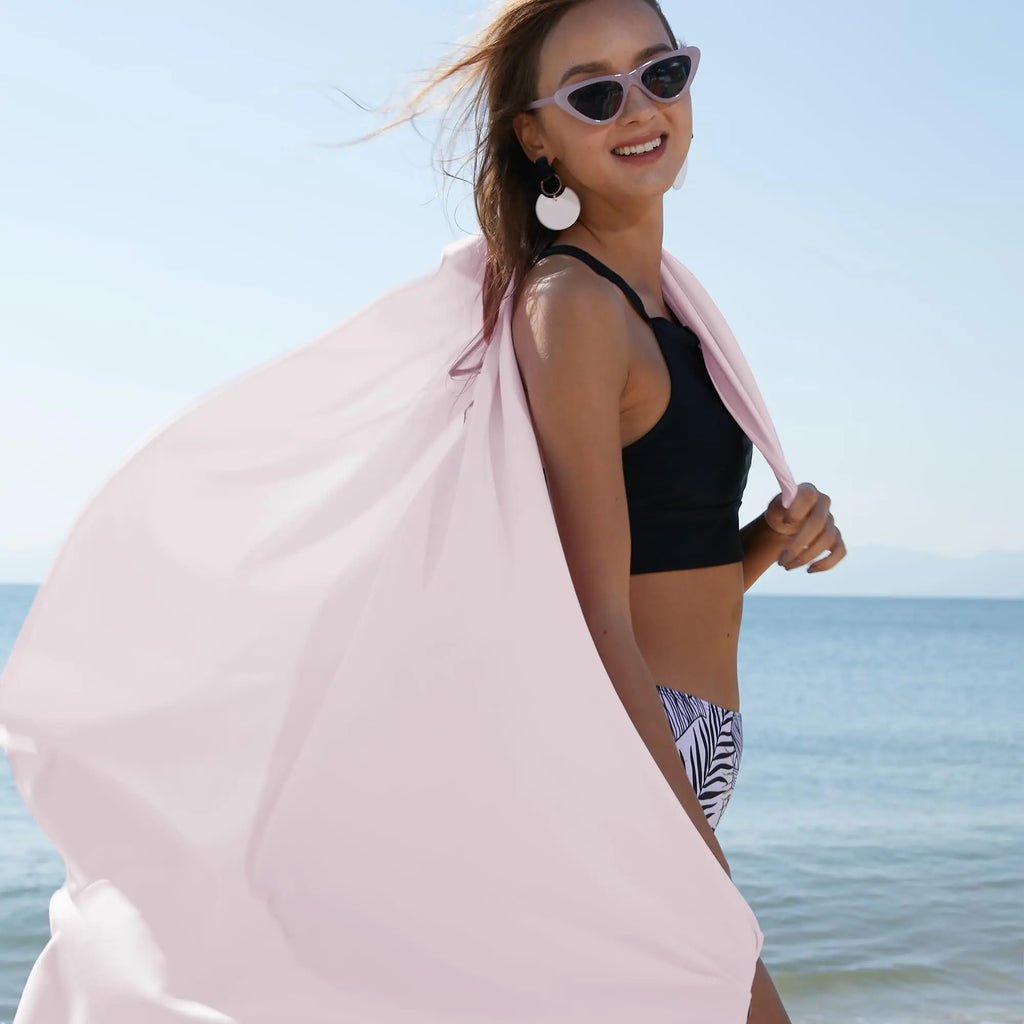 Woman wearing a black bikini and cat-eye sunglasses, holding a large, flowing light pink beach sarong by the sea.