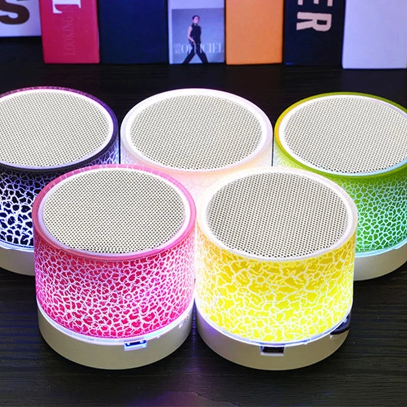 Five portable mini wireless speakers with crackle-effect LED light bases in pink, yellow, purple, white, and green.