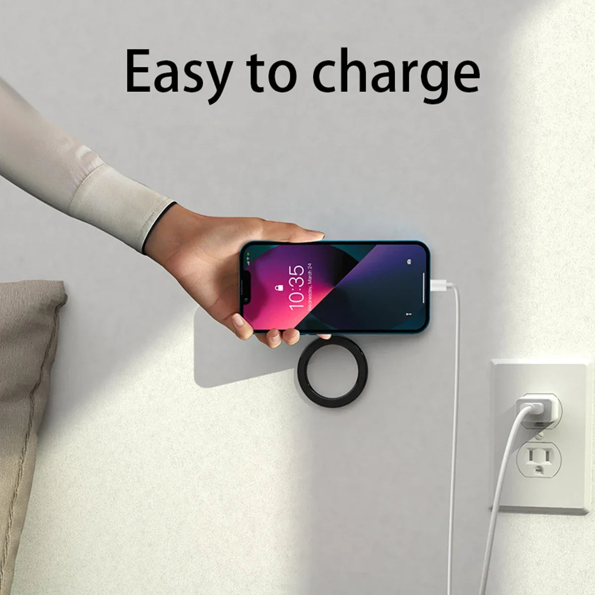 A hand holds a smartphone against a black, circular, wall-mounted magnetic wireless charger connected to a power outlet.