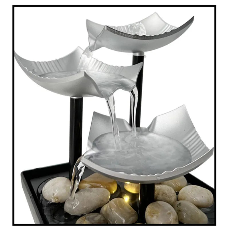 Three-tier silver tabletop fountain with black stands, water cascading onto decorative pebbles.