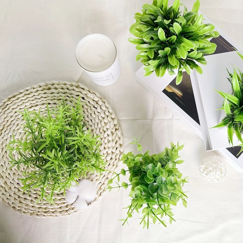 Top-down view of three small artificial green plants in white pots, arranged with a woven placemat and white decor.