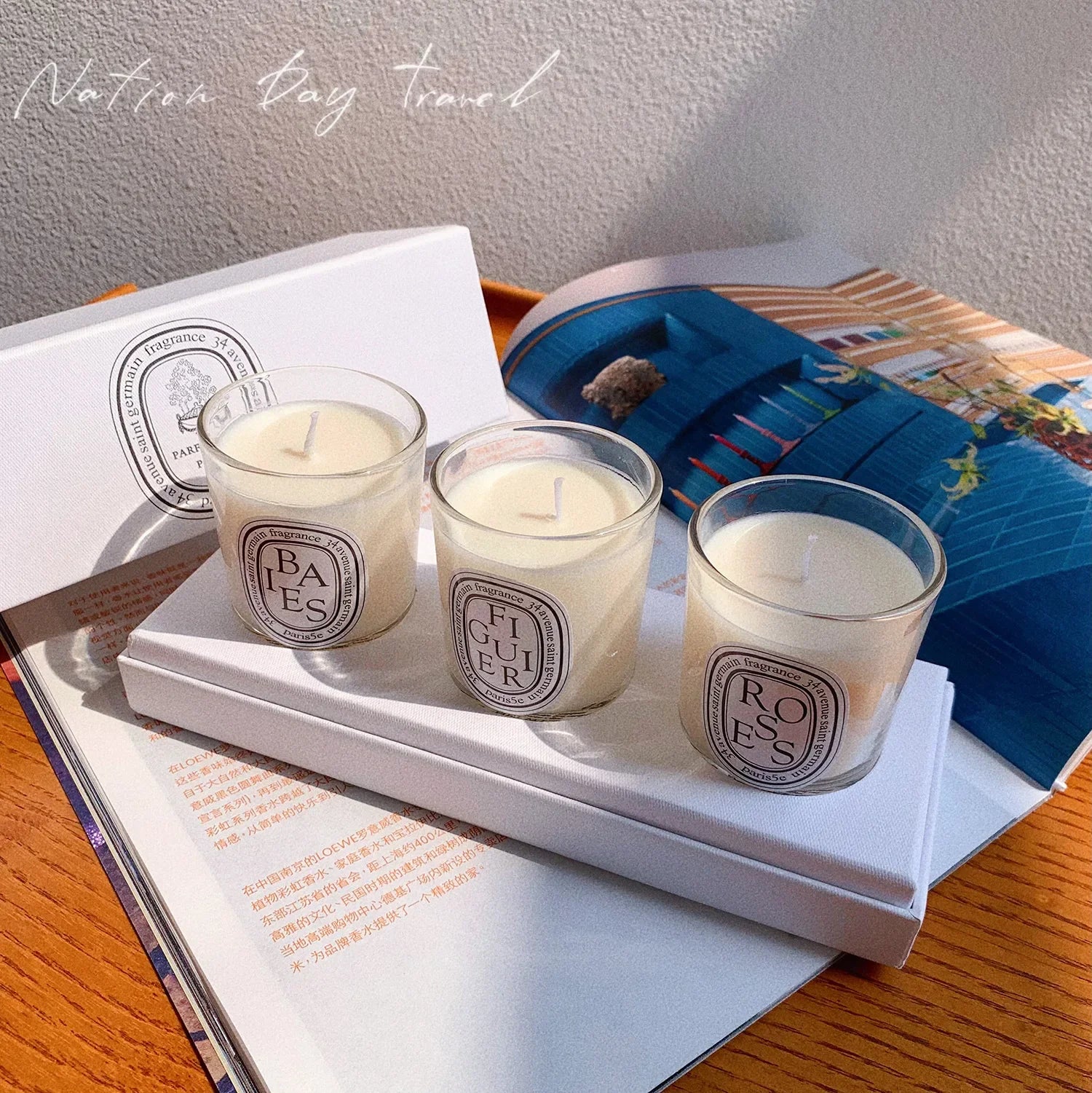 Three small scented candles in clear glass jars with cream wax and oval labels, displayed on a white box and magazine.