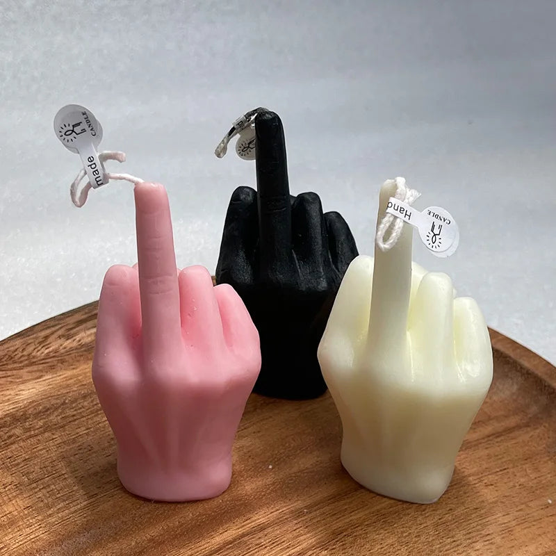 Three candles in pink, black, and cream, shaped like a hand giving a middle finger, sitting on a wooden tray.