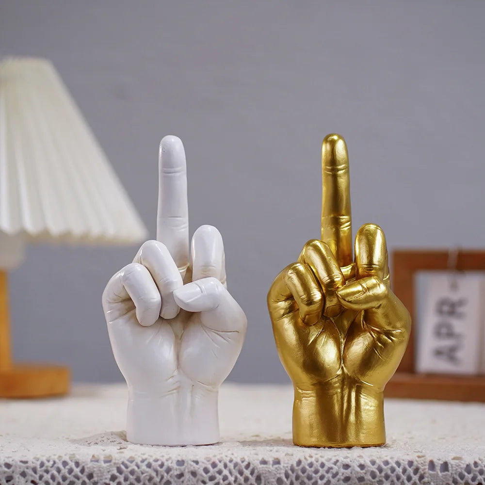 Two desk sculptures of hands in a middle-finger gesture, one in matte white and one in metallic gold finish.