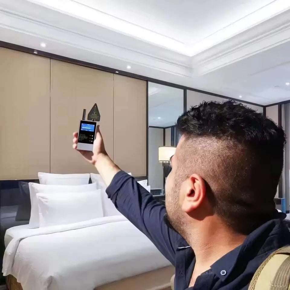 A person holds a compact, silver and black wireless signal detector with a digital screen in a modern hotel room.