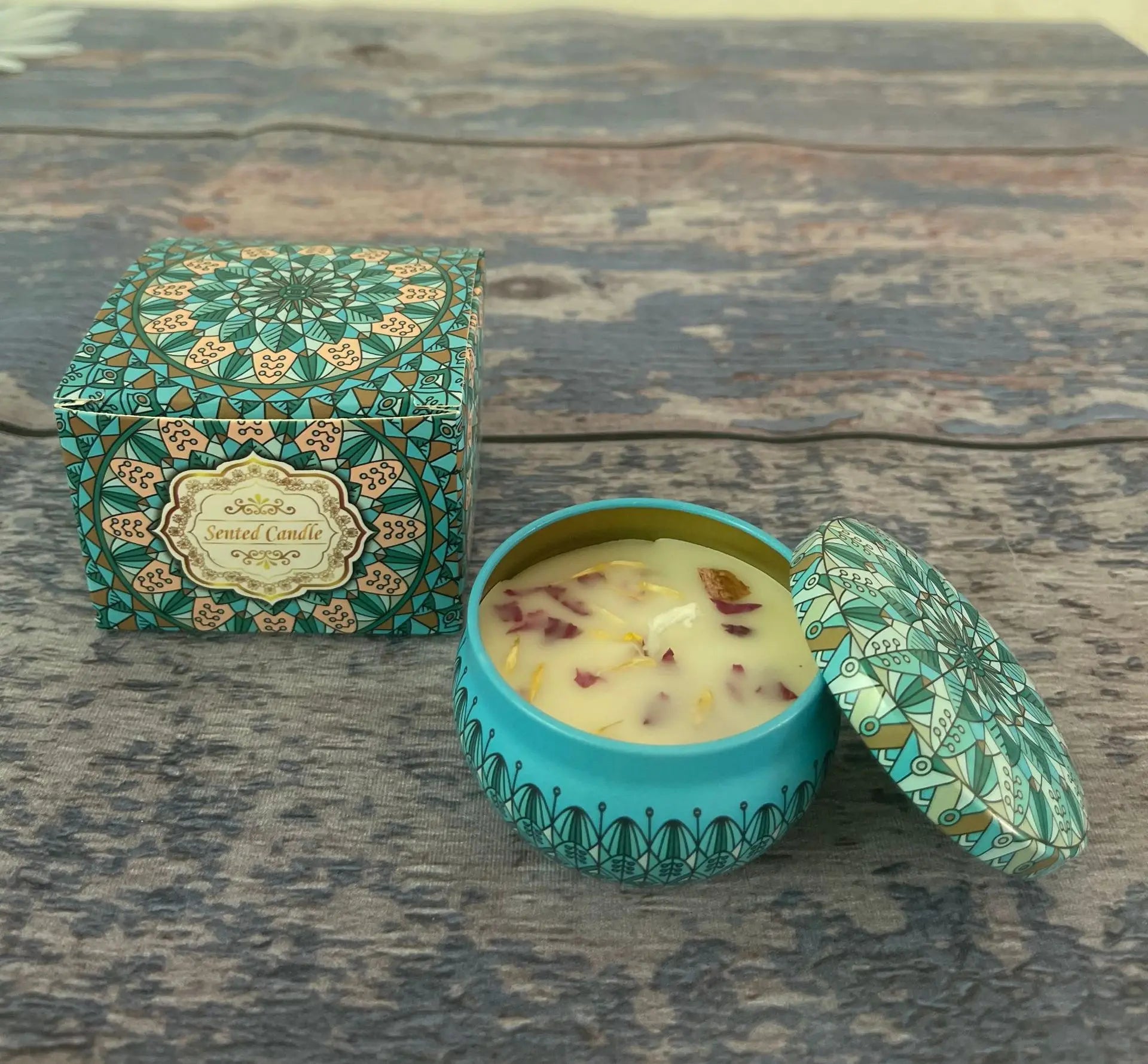 Scented candle in a teal tin with patterned lid, topped with flower petals, and matching floral-print gift box.