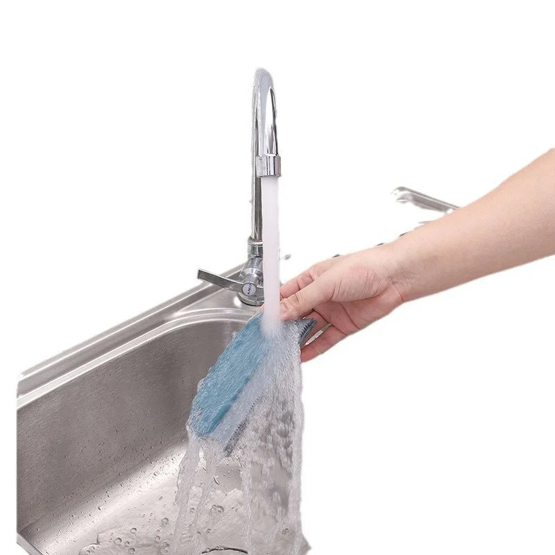 A hand rinsing a textured, grey-blue rectangular cleaning brush under a kitchen tap over a stainless steel sink.
