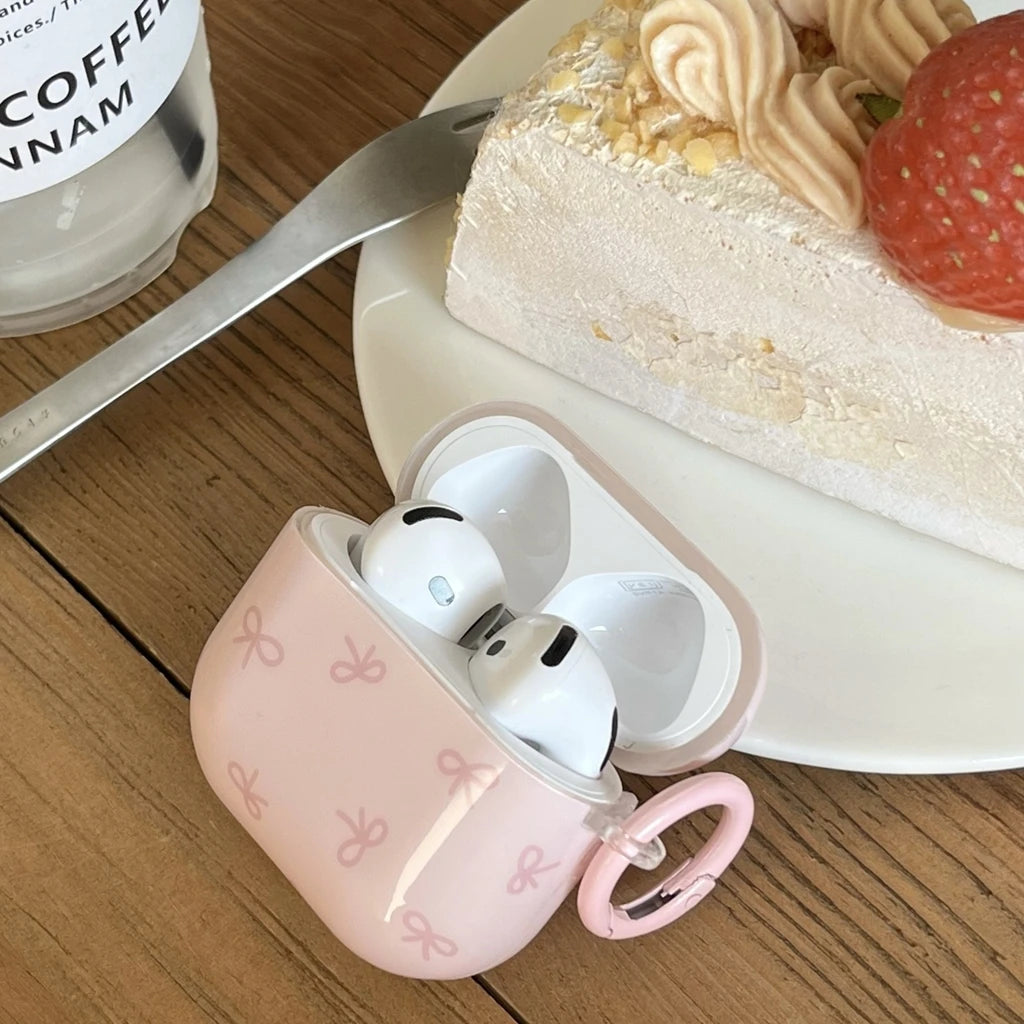 Glossy pink AirPods Pro case with a white bow pattern and a matching ring clip, displayed next to a dessert.