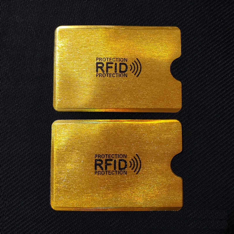 Two rectangular gold-colored metal RFID protection cards with a thumb notch, isolated on a black background.