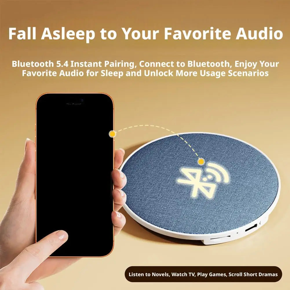 A hand holding a smartphone next to a round, fabric-covered blue wireless Bluetooth under-pillow sleep speaker.