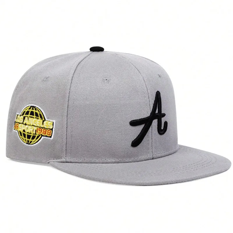 Grey baseball cap with a black letter 'A' embroidery and a yellow globe logo on the side. Flat peak hip-hop style.