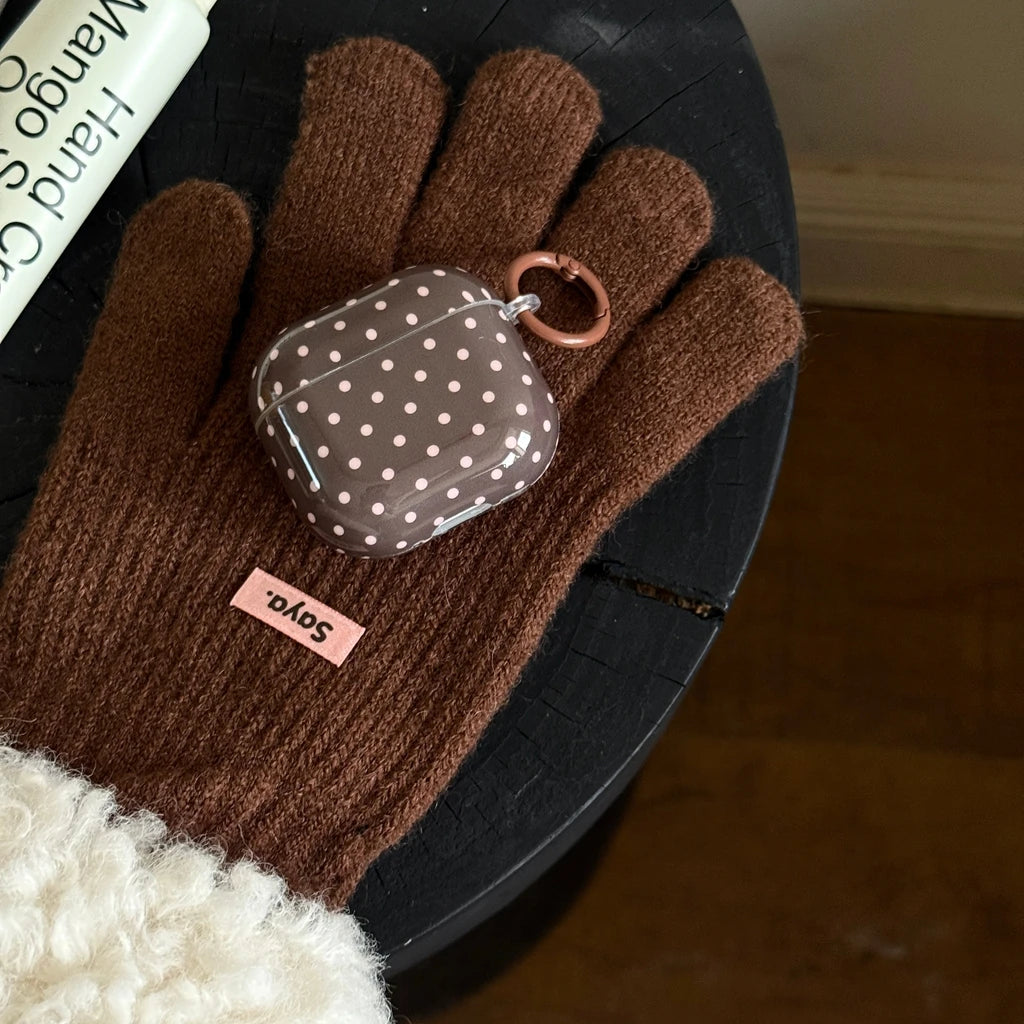Brown polka-dot AirPods case in hard plastic with a matching ring attachment, resting on a brown wool glove.