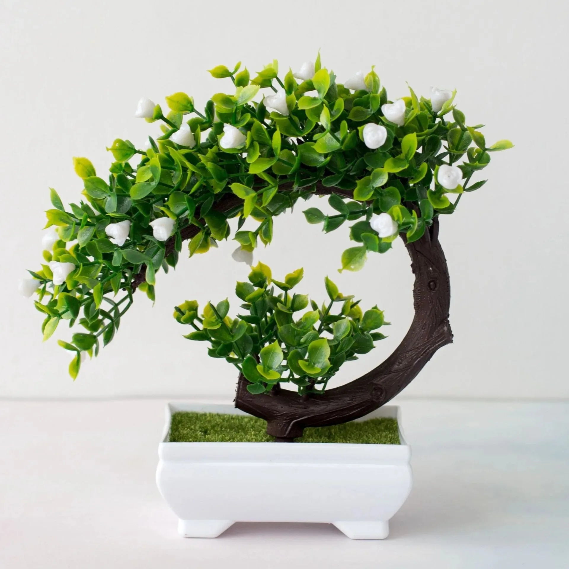 Artificial bonsai tree with green leaves and small white flowers in a curved shape, potted in a white rectangular vase.