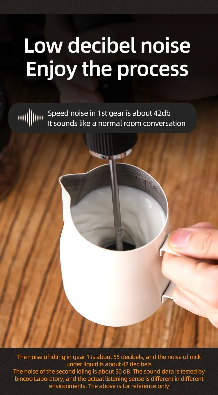 A handheld black milk frother whisking milk in a white stainless steel pitcher on a wooden surface.