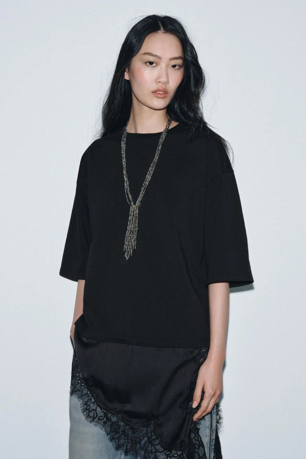 Model wearing a black oversized t-shirt, paired with a black lace-trimmed skirt over light-wash jeans and a long necklace.