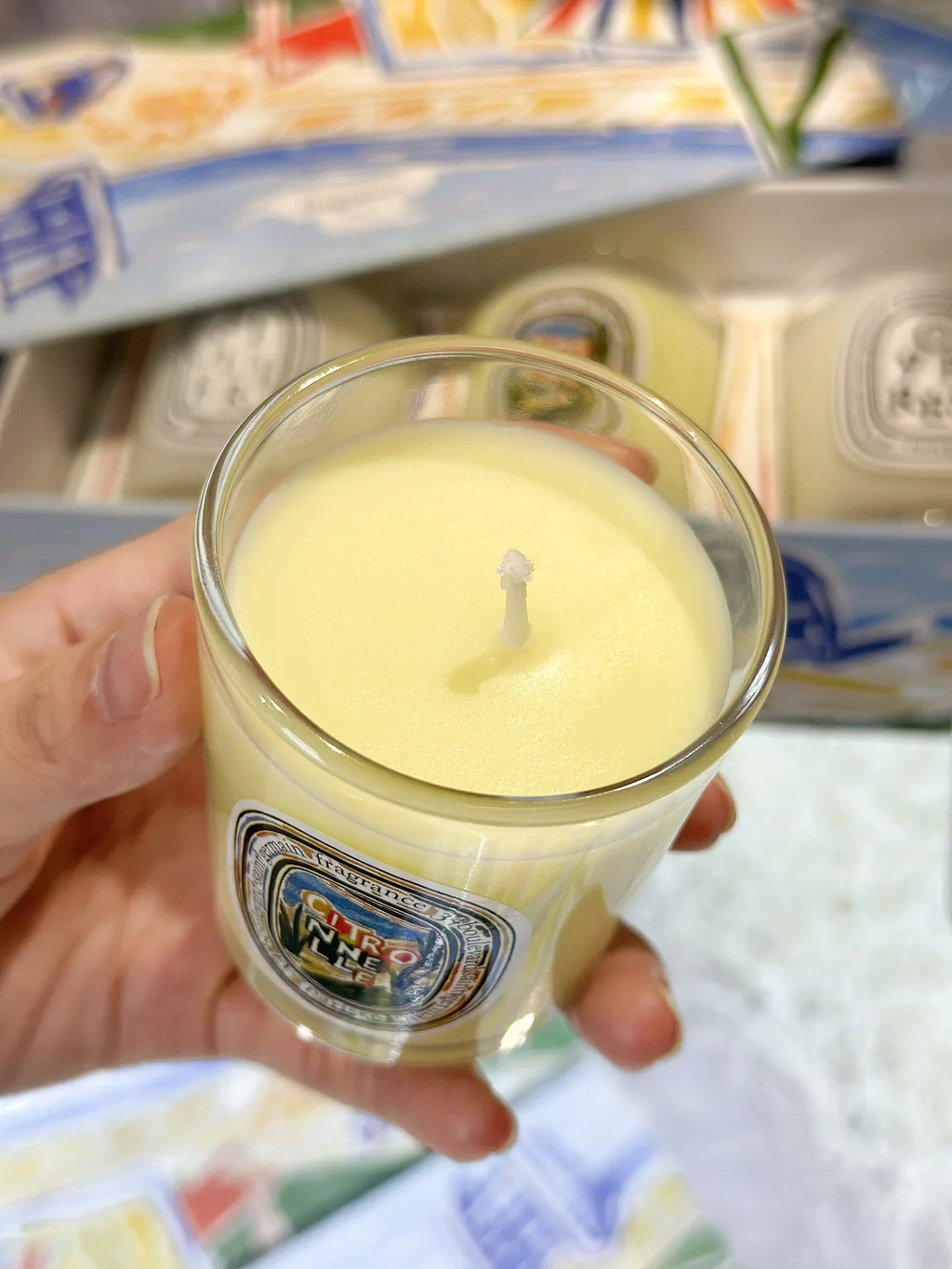 Hand holding a small, clear glass candle with pale yellow wax and a centered wick, with a decorative label.