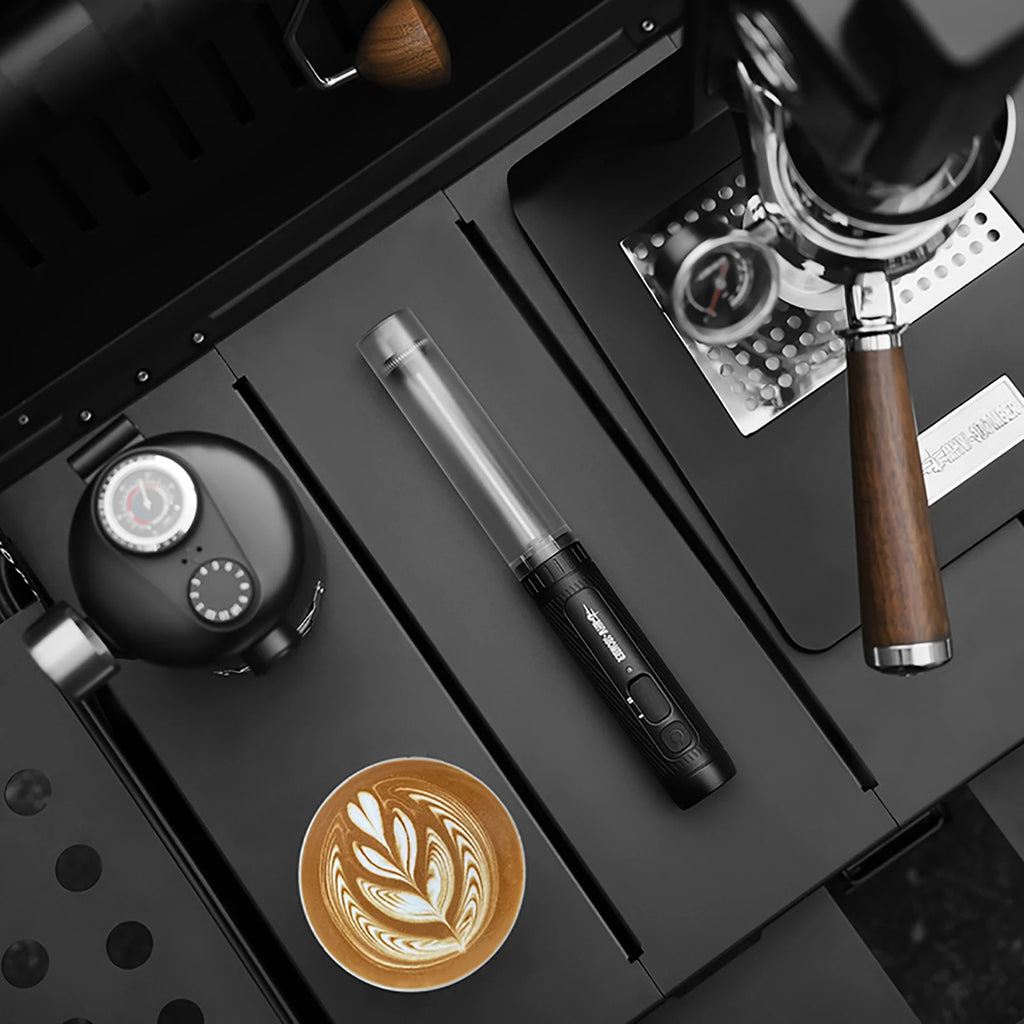 Black handheld milk frother with clear tube resting on a dark tray next to coffee equipment and latte art.