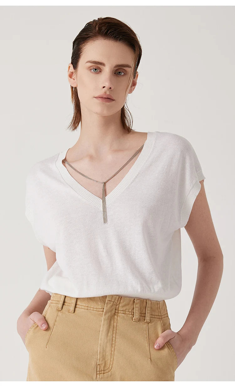 White short-sleeved V-neck top with a delicate silver chain detail at the neckline, worn with tan trousers.