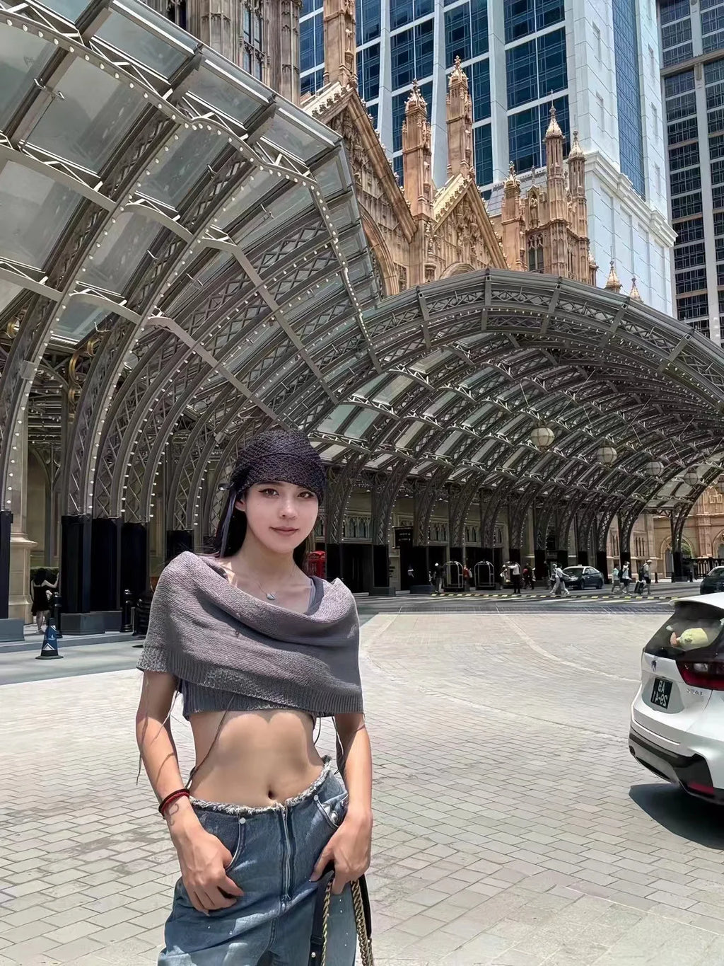 A woman wearing a dark grey, crochet-style knit hat with ribbon ties, posed in front of a grand arched building.