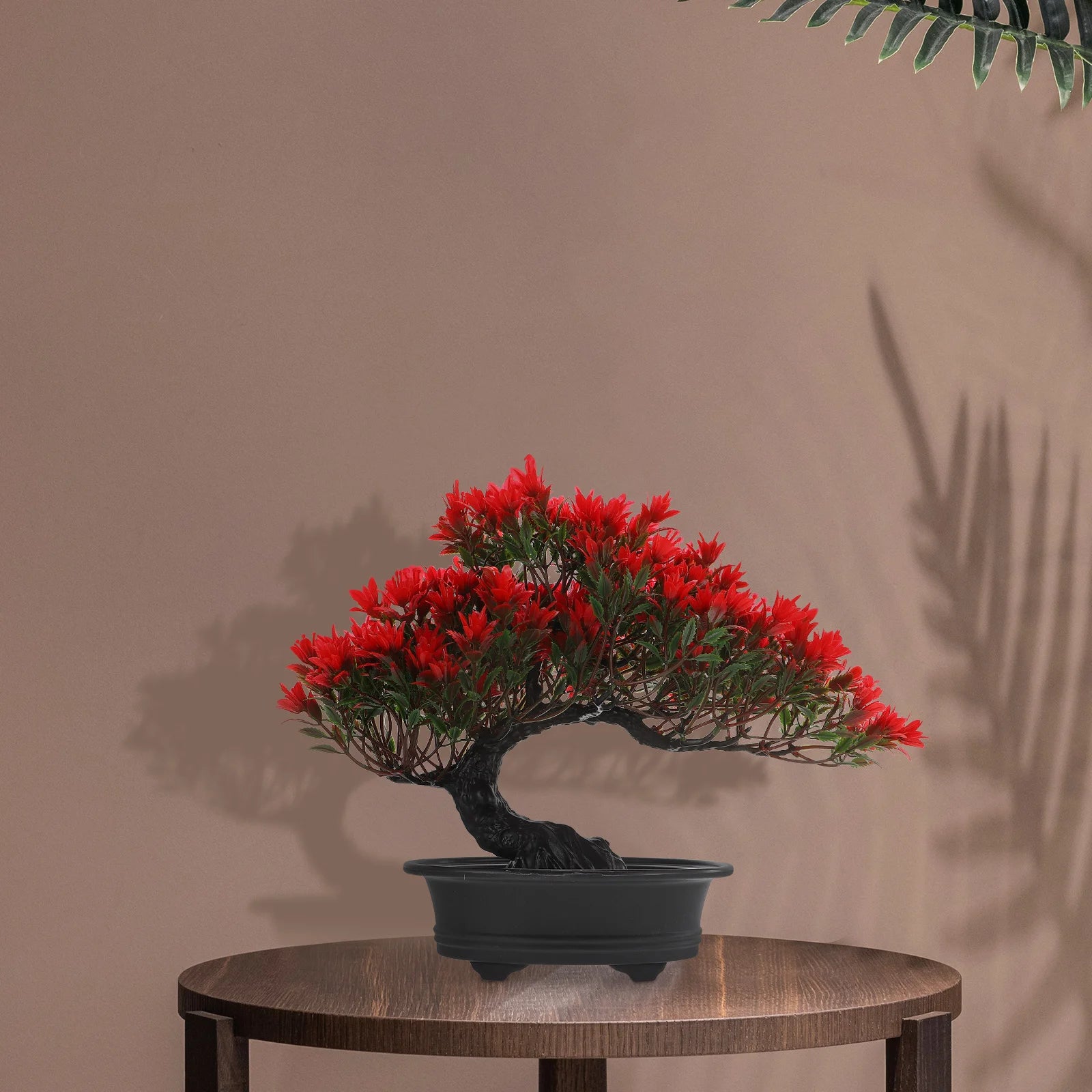 Artificial bonsai tree with vibrant red leaves in a sleek black pot, sitting on a wooden stool against a beige wall.