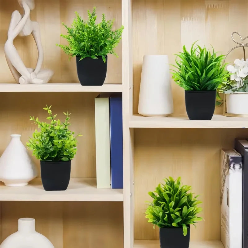 Four artificial green plants in small black pots displayed on a light wooden bookshelf with decor items and books.