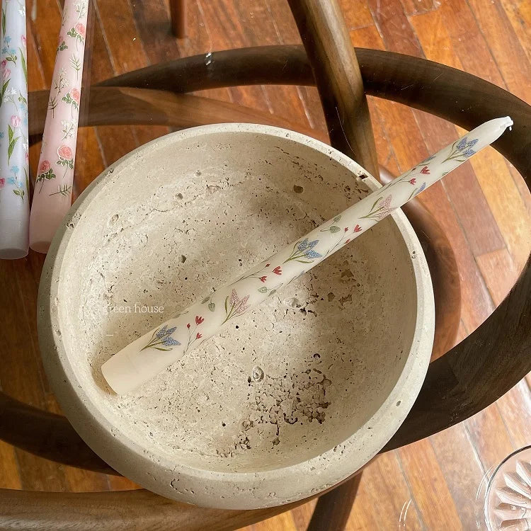Elegant taper candle in cream with a delicate blue and pink floral pattern, resting in a rustic stone bowl.