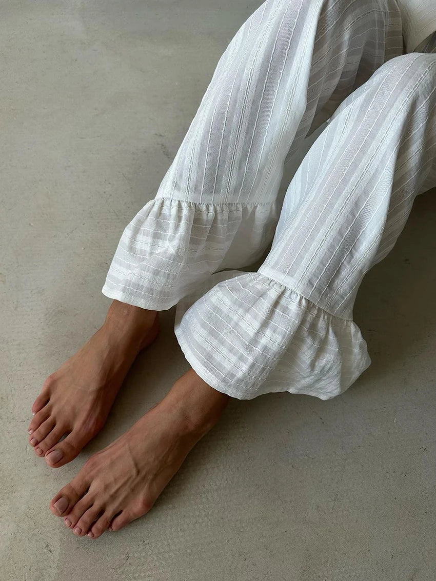 White cotton trousers with a textured stripe pattern and ruffled hems, worn by a person sitting on a concrete floor.