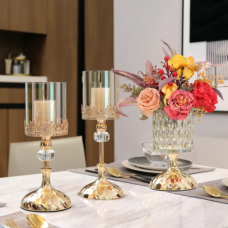 Two gold crystal candle holders with glass cylinders, one with white candles and another holding flowers.