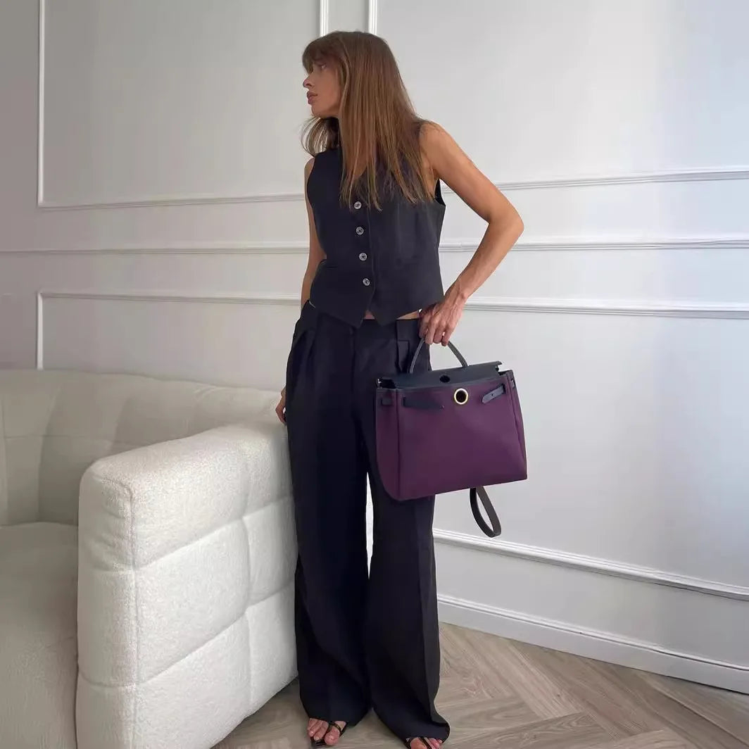 Woman in black waistcoat and trousers holding a structured, dark purple top-handle tote bag with gold hardware.